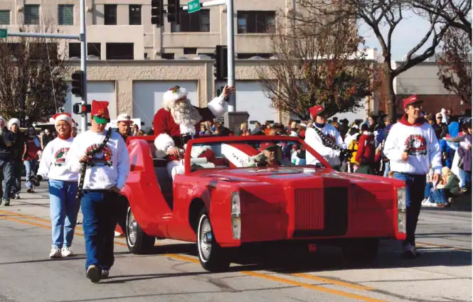 Santa Claus and parade participants in holiday costumes with a red vehicle in a community parade.