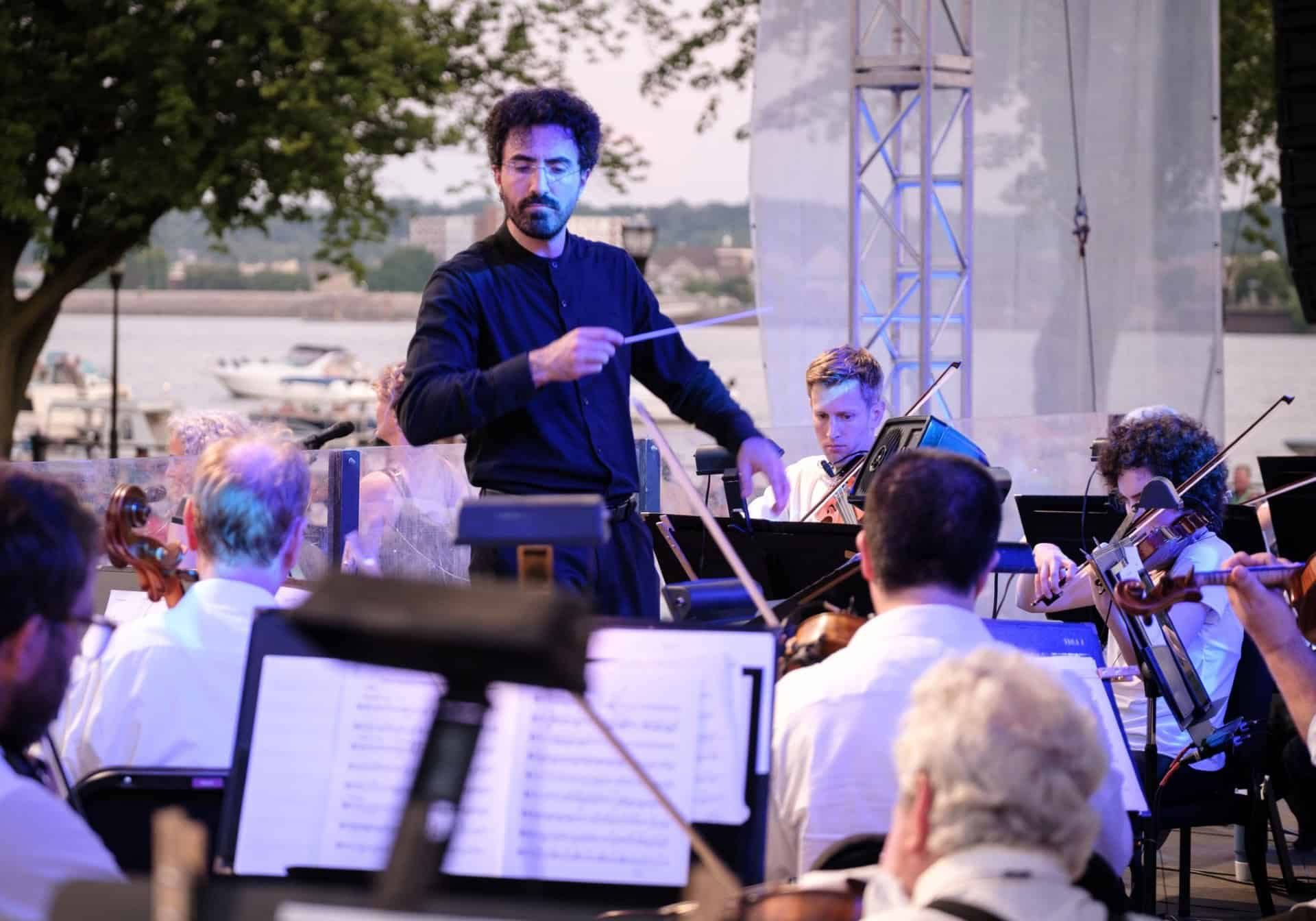Orchestra conductor leading musicians outdoors by the river at The Cultural Trust event.