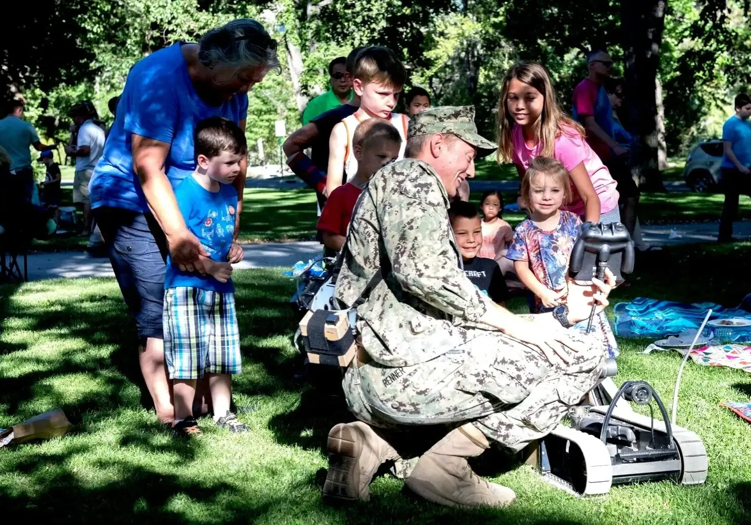 Military veteran demonstrating equipment to children outdoors in a park setting.